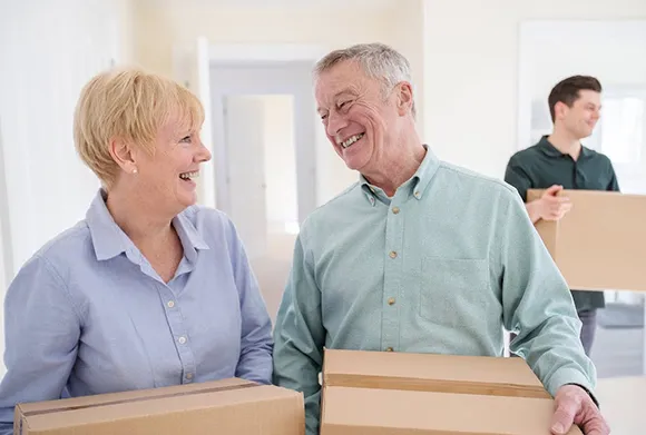 Happy elderly couple entering their new home, holding moving boxes, with their son in the background.